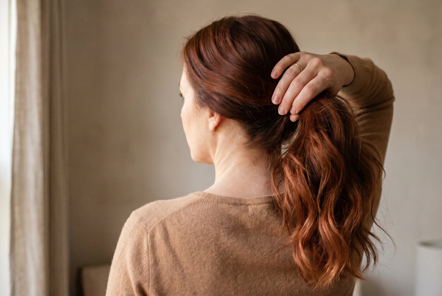 Woman with raw virgin auburn tape-in hair extensions in warm soft window light, photographed from behind
